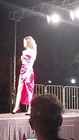 The video appears to be a fashion show. In the first frame, a woman in a pink dress walks down a runway. In the second frame, a woman in a white dress walks down the runway. In the third frame, a woman in a dark blue dress with lace detailing walks down the runway. In the fourth frame, two women are on the runway, one in a pink dress and one in a dark blue dress. The video is shot from the perspective of an audience member. There are stage lights and a metal barricade visible.