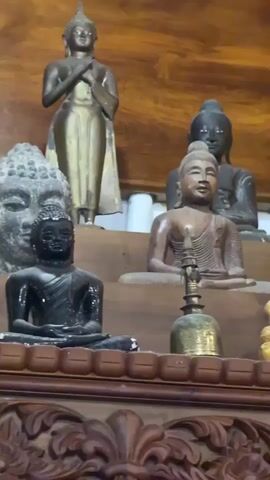 The video shows a collection of Buddha statues on a wooden shelf with ornate carvings. The statues vary in size, material, and color, including gold, bronze, and stone. Some are seated, while others are standing. A small brass bell sits in front of the statues. The background is a light brown wooden wall.