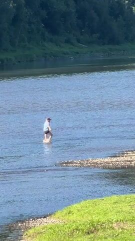 A person in a blue shirt and black shorts, wearing a pink hat, stands in a shallow river. The person is holding a white object, possibly a frisbee or disc, and appears to be throwing it. The river has a rocky bottom and the water is clear. In the background, a dense green forest lines the opposite bank of the river, and the sky is bright and sunny. The foreground shows a grassy bank with small stones.