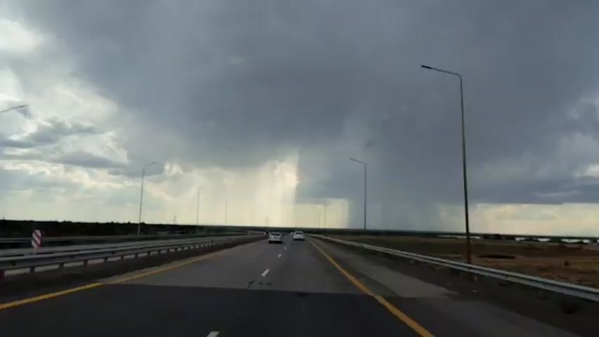 The video captures a perspective from a vehicle driving on a highway. The sky is filled with dark, heavy storm clouds from which streaks of rain are visible falling in the distance. Several cars are driving on the highway ahead and to the side. The road has multiple lanes and is bordered by metal guardrails and streetlights. The landscape to the sides of the highway appears to be dry, with some low vegetation and flat terrain extending towards a body of water or a distant plain. The overall atmosphere is one of an approaching or ongoing storm.