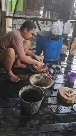 A person is squatting on a wet wooden porch, preparing fish. The person is wearing a tan t-shirt and orange flip-flops. They are holding a knife in their right hand and a fish in their left hand, which they are cutting on a large, round wooden cutting board. There are several other fish on the cutting board, along with some fish entrails. To the left of the cutting board, there is a black bucket that appears to contain fish guts. In front of that bucket is another large, rusty metal bucket. To the right of the cutting board, there is a small wooden cutting board with a single fish on it. Behind the person, there is a large blue plastic barrel, a white bottle with a pump dispenser, and some white plastic bags hanging from a rack. The porch has a railing and a dark, wet surface. The background shows some green structures and netting. The overall environment appears to be outdoors, possibly near water.