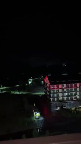 A video shows a nighttime aerial view of a resort complex with a main hotel building and several smaller lodges. The buildings are illuminated with red and green lights. A parking lot with several cars is visible in the foreground. The ground appears wet, suggesting recent rain. The sky is dark.