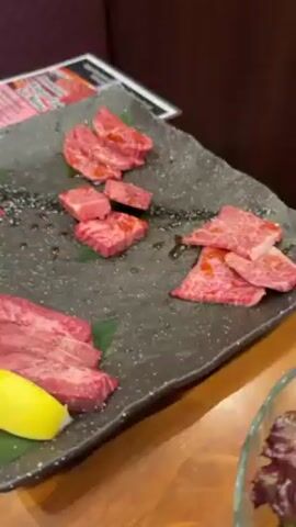 The video shows a plate of raw meat, possibly at a Japanese barbecue restaurant.  The meat is arranged in several portions on a speckled, dark gray plate.  The cuts vary in shape and size, some are sliced and some are cubed. Some are topped with small green leaves, a few drops of dark sauce, and one portion has a lemon wedge beside it. The plate sits on a wooden table, with a blurry menu partially visible behind the plate. A bowl of salad is slightly visible on the right side of the plate.  The background shows a booth with a maroon-patterned back.