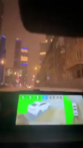 The view from inside a car moving down a city street at night is captured in this video. High-rise buildings and skyscrapers are illuminated in the background. Raindrops can be seen on the windshield. The car’s dashboard is visible with an infotainment system displaying music controls. The music playing is titled "Vmeste My" by AMCHI Band.
