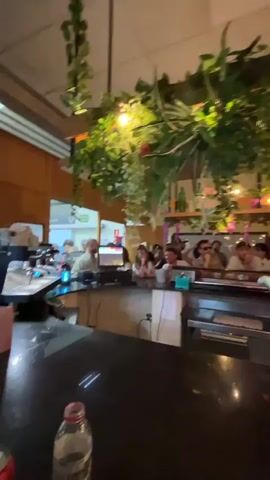 The video opens with a view of a bar area decorated with hanging plants and lights. Several people are visible standing or sitting at the bar, and some appear to be interacting with staff behind the counter. The camera then pans to show a group of people standing near an entrance, some of whom are looking at their phones. The scene appears to be a lively indoor environment, possibly a restaurant or bar.
