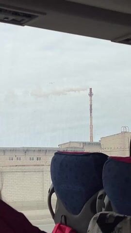 A person is riding in a vehicle, possibly a bus, and films a view of an industrial area through the window. The scene shows a tall smokestack emitting smoke, with brick buildings and a large red crane visible in the background. The sky is overcast. The vehicle's interior is visible, with blue seats in the foreground.