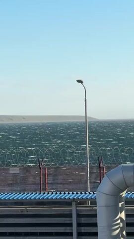 The media file captures a windswept seascape with industrial elements in the foreground. The choppy, dark green water is indicative of a strong breeze, with whitecaps dotting the surface. Beyond the water, a distant, rolling landmass is visible under a clear, light blue sky.

In the middle ground, a tall lamppost stands sentinel, topped with a single light fixture. Below it, a fence constructed of wire coils and red-capped metal posts acts as a barrier. Below the fence, a section of a structure with a blue, corrugated roof and grey metal siding is visible. To the right of the frame, a large, curved metal pipe, likely part of an industrial complex, angles upwards.

The scene appears to be set during daylight, possibly late morning or afternoon, given the bright sky and clear visibility. The weather is windy, as evidenced by the turbulent water. The presence of the industrial infrastructure and the barbed wire fence suggest a secured or functional area, possibly a port or a processing plant. The vastness of the water and the distant land create a sense of openness, contrasting with the man-made barriers. No people are visible in the frame, and there is no discernible text. The overall mood is one of industrial activity in a natural, yet potentially harsh, environment.