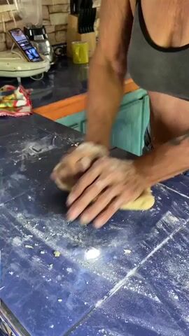 The video shows a person kneading dough on a blue tiled countertop. The person's hands are covered in flour, and they are using their palms and fingers to press and stretch the dough. The dough appears to be light yellow and has a slightly sticky texture. There is also some loose flour scattered on the countertop. In the background, a kitchen counter is visible with a white appliance, a phone, a stack of colorful napkins, and a wooden knife block with knives. A cabinet with light blue doors is also visible behind the countertop.
