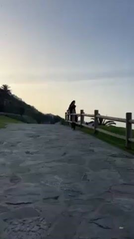 The video shows a black cat walking towards the camera on a stone pathway. In the background, a person stands by a wooden fence. The area appears to be outdoors, with some greenery and trees visible on a hillside. The sky is overcast. The cat meows as it approaches.