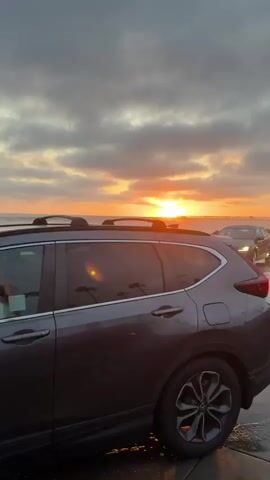 A gray SUV is parked in the foreground facing right. The ocean is in the background, with waves crashing against a concrete barrier. The sun is setting, casting an orange glow across the sky and reflecting on the wet road. A dark sedan is driving on the road, heading towards the sunset. A person on a bicycle is riding on the road behind the sedan. The sky is cloudy with streaks of orange and yellow from the sunset.