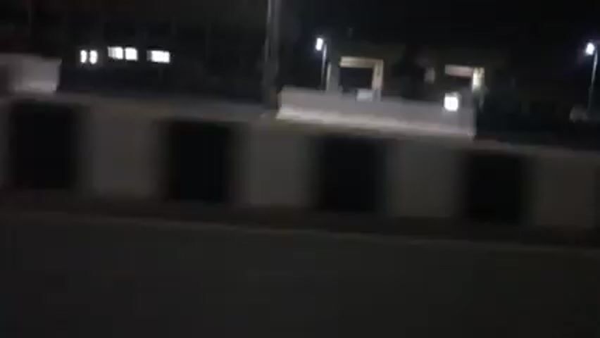 The video shows a night scene from a moving vehicle. The camera is aimed toward a roadside area featuring a building with illuminated sections. There are streetlights providing illumination along the road and around the building. The road is separated from the roadside area by a barrier. As the vehicle moves, a police car with flashing red and blue lights becomes visible. The surroundings are dark.