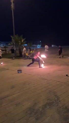 A fire dancer is performing in front of an audience at night on a wooden boardwalk. The fire dancer is holding two flaming props and spinning them, creating arcs of light. The audience members are seated and standing behind the fire dancer, watching the performance. In the background, there are palm trees, beach chairs, and a dark sky. The scene is lit by the flames of the props and some artificial lighting from poles.
