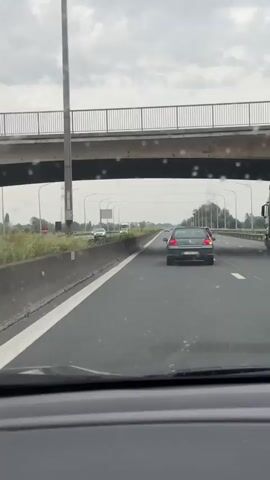 The video shows a car driving on a highway on a cloudy, rainy day. The view is from inside the car, looking out through the windshield, which is covered in raindrops. Other cars are visible on the highway, moving in the same direction. The highway has multiple lanes and is bordered by a concrete barrier and green vegetation on the side. Overhead, a bridge crosses the highway. Tall lamp posts line the side of the road, and a power line tower is visible in the distance.