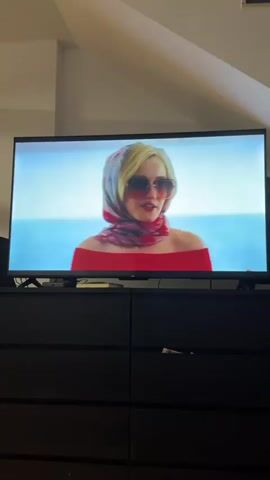 A television displays a scene from a movie or show. Four people are on what appears to be a boat. On the left, a woman with blonde hair wears a colorful, patterned robe and sunglasses. In the center, a woman with blonde hair wears a red dress, a headscarf, and sunglasses, holding a glass of champagne and a handbag. To her right, a woman with dark hair wears a blue off-the-shoulder dress with a colorful pattern and holds a drink. On the far right, a person with dark hair and a patterned shirt is partially visible. The people are interacting, with the woman in the red dress speaking to the woman in the blue dress. The background shows the ocean and a blue sky. The television is placed on a dark dresser with several drawers.