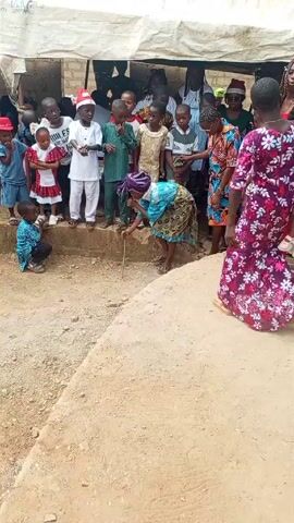 A woman, dressed in a bright blue and patterned dress and headwrap, bends over, holding a microphone to the ground in front of a small group of children. One child sits on the ground watching her. The other children are standing on a raised area covered in tan-colored soil. Some of the children are wearing festive clothing. Behind the children is a structure covered with a white tarp, and several adults are standing there. The ground in front of the woman and the children is rough and mostly dirt.