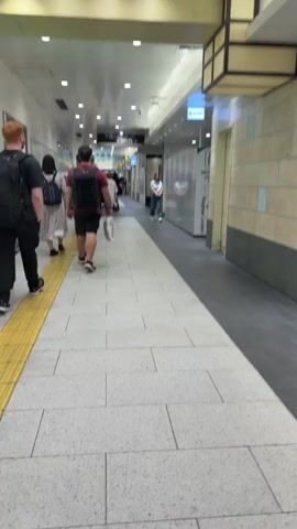 A group of people are walking down a paved hallway. The hallway has white tile floors, a yellow tactile paving strip on the left side, and fluorescent lighting from the ceiling. On the right side of the hallway are lockers labeled "Baggage Lockers." Several people are walking away from the camera, and one woman is walking towards the camera. The people are dressed in casual clothing. Some are carrying backpacks or bags. The environment appears to be a train station or airport.