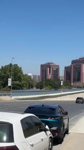 This is a vertical, eye-level, handheld shot of an urban street scene in Bakırköy, Turkey. The day is bright and clear, with a vibrant blue sky and ample sunlight, suggesting it is midday on a warm day. The foreground features two cars parked along a sidewalk, one white and one dark green. Beyond them, a multi-lane road curves into the distance. A white bus with a Turkish flag decal on its rear travels on the road. In the mid-ground, a concrete barrier separates the road from a green, grassy slope. In the background, several modern, large, multi-story buildings dominate the skyline. These buildings appear to be commercial or office spaces, with a symmetrical architectural style and dark-colored facades. Several lampposts line the sidewalk and road, with banners hanging from some of them. One banner prominently displays colorful, abstract squiggles and the Turkish text "tatil budur." Another banner, further down the road, also appears to have text on it. The overall impression is a busy, functional urban environment under a clear, sunny sky. The presence of cars and buildings indicates a developed area, likely in a city.