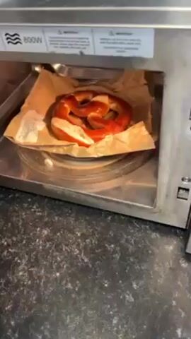 A hand opens the door of a microwave oven with a pretzel inside. The pretzel rests on a piece of parchment paper. The hand rotates the timer dial to set the microwave to 15 seconds. The microwave starts running.