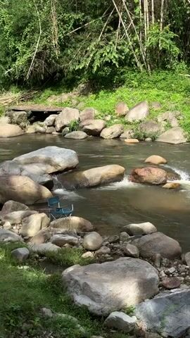 The video shows a flowing river surrounded by large rocks and lush green foliage. Two blue camping chairs are placed on the rocky bank of the river. The water is clear and a small waterfall is visible where the river narrows between rocks. The sun is shining, casting dappled light on the scene. The overall atmosphere is serene and natural.