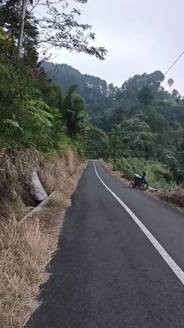 The video shows a winding road in a mountainous, rural area. The road is paved with asphalt and has a white line down the center. On the left side of the road, there is a steep, vegetated slope with dry grass and some green foliage. On the right side of the road, a motorcycle is parked. Beyond the motorcycle, the terrain slopes down into a valley filled with lush green trees and vegetation. In the distance, more rolling hills covered in trees are visible under a cloudy sky. The overall impression is one of natural beauty and isolation.