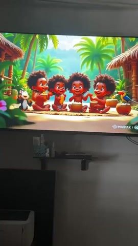 The video shows a television screen displaying an animated scene. Four cartoon babies with dark skin and curly hair are sitting in a row on what appears to be a sandy area. They are surrounded by lush greenery, palm trees, and thatched huts, suggesting a tropical environment. A monkey is visible to the left of the babies, and a bird is perched on a fruit to the right. The babies are all looking forward and seem to be interacting, with some holding bowls. The lower right corner of the screen displays "MINIMAX" and a logo, indicating the channel. Below the television screen, a shelf holds various items, including bottles and decorative objects.