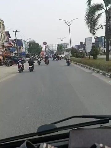 The video shows a view from a vehicle moving down a paved road. The road is lined with buildings on the left and a grassy median with a curb on the right. Several motorcycles and a few cars are visible on the road, moving in the same direction as the camera. The sky is overcast. The buildings on the left appear to be businesses, with signs for "Alfamart" and "BAKSO". There are also power lines and streetlights visible. The overall impression is a busy urban or suburban street scene.