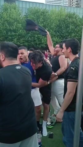This is a video taken from a crowd of people outdoors. The camera pans across the faces of several individuals, some of whom are shouting and cheering. The people are dressed in casual clothing, and some are wearing sports jerseys. In the background, there are trees and buildings, suggesting an urban setting. The overall atmosphere appears to be one of excitement and celebration.