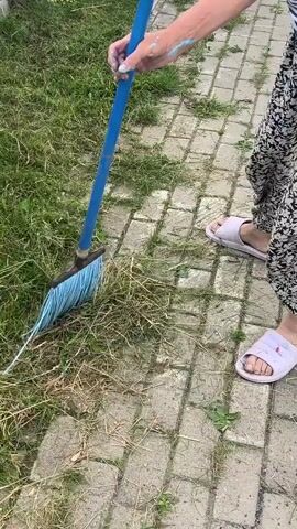 The video shows a person's legs and arms as they use a blue rake with a blue head to gather grass and debris from a brick pathway. The person is wearing black and white floral pants and white slippers. The environment appears to be outdoors, with green grass and a paved walkway.