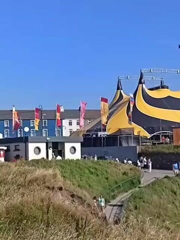 This is a video of a large, black and yellow striped circus tent set up on a grassy hill overlooking a body of water. A group of people are gathered around the tent, some entering and exiting. A ramp leads up to the tent entrance. In the background, there are buildings and trees. The sky is clear and blue.