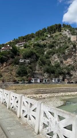 The video opens with a wide shot from a bridge, looking towards a steep, green hillside under a bright blue sky with scattered white clouds. The bridge's white concrete railing, featuring a distinctive geometric pattern, is prominent in the foreground. Below the bridge, a light green river flows, and beyond it, a road runs along the base of the hill where a few cars are parked. The hillside is densely covered in trees and shrubs, with several white-roofed buildings scattered across its slopes, leading up to what appears to be a stone wall or ruins at the summit.

As the camera pans slightly, a white drone (resembling a DJI Mini series) enters the frame, flying in the middle distance against the clear blue sky. The drone is seen from below, with its propellers spinning as it hovers. The perspective then shifts to show the drone from the side, with the hillside and its buildings in the background, offering a view of the drone's body and camera. The video captures the drone in stable flight, with the scenic Albanian landscape as its backdrop.