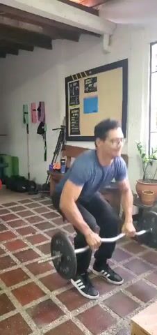 A man is filmed performing a multi-part exercise sequence indoors in El Papelón, Venezuela. He is wearing a blue t-shirt, dark sweatpants, and black and white Adidas sneakers. He has short dark hair, glasses, and a mustache.

The video opens with the man sitting on a reddish-brown tiled floor, holding an EZ curl barbell with weights loaded on each end. He appears focused, with a slight smile. The weights are marked "10 COLD," indicating 10 kilograms. From this seated position, he smoothly transitions to lying on his back to perform a floor press. After completing the press, he sits up, bringing the barbell back to his lap, and then stands up, holding the barbell in front of him. He concludes the sequence by performing an overhead press, extending his arms fully above his head, maintaining a determined expression throughout the movement.

The setting appears to be a home gym or a small, well-equipped fitness studio. The walls are white, and a wooden beam structure is visible on the ceiling. In the background, various pieces of gym equipment are visible: a wooden bench holding a red kettlebell, a dark water bottle, and additional weight plates. Above the bench, a large bulletin board with a dark frame and yellow accents displays several posters or papers. To the left, resistance bands in various colors (pink, green, purple) hang on the wall, and a tripod stands on the floor next to other dark gym equipment. A window on the right side provides natural light, and a potted plant rests on a small wooden stool. The man's movements are controlled and fluid, suggesting a regular fitness routine.