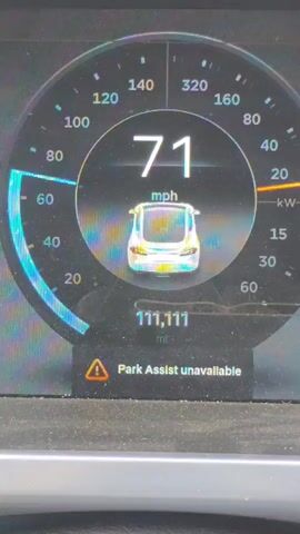 The video begins by showing the dashboard of a car. The speedometer displays a speed of 71 mph, and the odometer shows 111,111 miles. A warning message states "Park Assist unavailable." The perspective then shifts to show the road ahead from inside the car. A pickup truck with a bike rack carrying bicycles is in the left lane, and a silver car is in the right lane, both ahead of the viewer's vehicle. The road is paved, lined with trees on both sides, and the sky is blue with some clouds. The vehicles are traveling on a highway under bright daylight conditions.