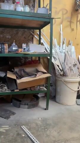 The video shows a cluttered workshop or storage area. On the left is a green metal shelving unit with various items on its shelves, including cans, bottles, and spools of wire. Below the shelves is a cardboard box overflowing with what appear to be metal parts and a large metal ring. To the right of the shelves, there are two large buckets filled with pieces of wood or other materials. A broom with a blue head leans against one of the buckets. The wall behind the items is painted yellow and has some graffiti or markings on it. A calendar hangs on the wall. Further to the right, there is a propane tank, a generator, and various pieces of cardboard and metal scraps on the floor. In the foreground, the concrete floor is visible with a few small items scattered on it. The lighting suggests it is daytime with sunlight streaming in from the right. There are no people visible in the video.