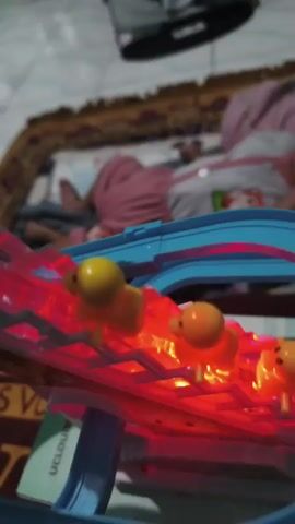 The video shows a toy duck slide with several yellow toy ducks ascending a moving staircase. The staircase is illuminated with red lights. In the background, a person is lying on a bed, partially visible. The toy includes a blue track and a yellow duck-themed sign with the words "Small Duck". The lighting is dim, suggesting an indoor environment.
