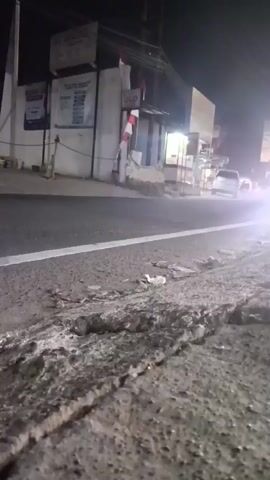 A low-angle shot captures a cracked concrete roadside at night. Beyond the road, a building with "UGD" signage is illuminated. A white car is visible in the distance, approaching a bright light source. A motorcycle with two riders passes by the building. The scene appears to be a street in a town or city at night.