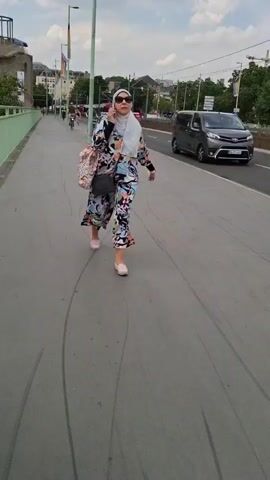 A woman wearing a hijab, sunglasses, and a colorful dress walks on a paved path while talking on a cell phone. She carries a large tote bag over her left shoulder and a small black purse across her chest. A dark-colored van drives by on the road to her right. A cyclist wearing a helmet and a backpack rides a bicycle towards the camera on the same path. The environment appears to be an urban setting with buildings and trees visible in the background, and a cloudy sky above.