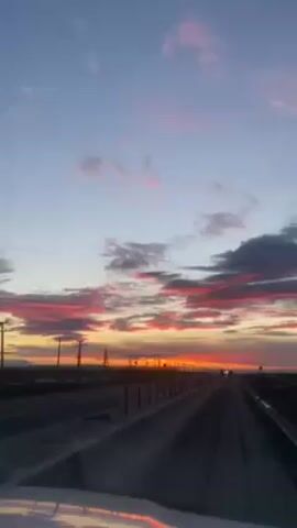 The video shows a view from inside a moving vehicle, looking out at a highway with a vibrant sunset in the background. The sky is a mix of light blue and orange with streaks of pink and dark clouds. Utility poles line the roadside. Cars are driving in the distance on the highway. The road has a dividing barrier along it. The reflection of the sky is seen on the vehicle's hood.