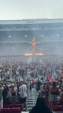 The video shows a concert performance in a large stadium. A towering golden statue of a person stands on a stage. The statue is illuminated with orange lights and smoke. A large crowd of people is gathered in front of the stage, many holding up their phones to record the event. The stadium seats are filled with spectators. The overall atmosphere appears to be energetic and exciting.