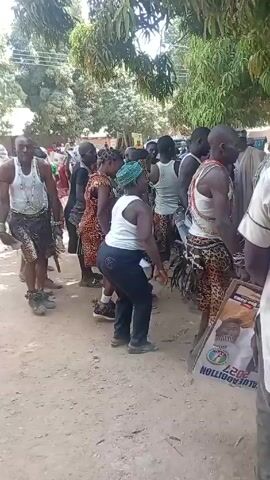 The video depicts a group of people dancing outdoors during the day. The dancers are wearing various outfits, including tank tops, patterned shorts or skirts, and beaded necklaces. Some carry small wooden sticks. A few wear traditional-style leopard print clothing. A sign with the word "Vaccination" on it is visible. The crowd appears to be gathered under trees, and onlookers are filming the scene with their phones. The ground is covered with dirt and dust.