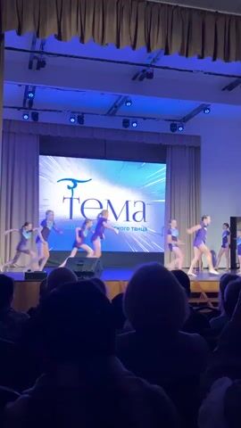 From a dark audience perspective, a stage is illuminated with blue light and contains a large LED screen showing "Tema, клуб акробатического танца." On the stage, about ten young dancers in purple outfits perform a dance routine. The scene is indoors, seemingly in a theater or auditorium. Some audience members are visible, a few holding up phones to record the performance. The overall mood is celebratory and focused on the performance.
