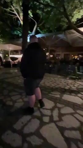 The video shows a person walking at night on a stone-paved surface, possibly a sidewalk. The person is wearing dark shorts, a dark jacket, and dark shoes. Trees and dim lighting surround the area, and there's a blurred glimpse of outdoor restaurant seating with tables and umbrellas.  A building with a neon sign is visible in the background. A second person appears briefly at the end of the video. Music plays in the background. The camera movement is shaky.