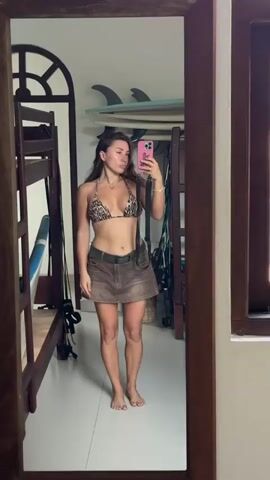 This media file features a young woman posing in a mirror, taking a selfie. She is wearing a leopard print bikini top and a short brown denim skirt. The setting appears to be an indoor space, possibly a bedroom or a dorm room, with a bunk bed visible to her left. Surfboards are stacked on shelves behind her, suggesting a beach or surf-oriented location. The lighting is bright, indicating daytime. The woman is making playful facial expressions and hand gestures, appearing confident and in good spirits. There is no visible text in the media file.