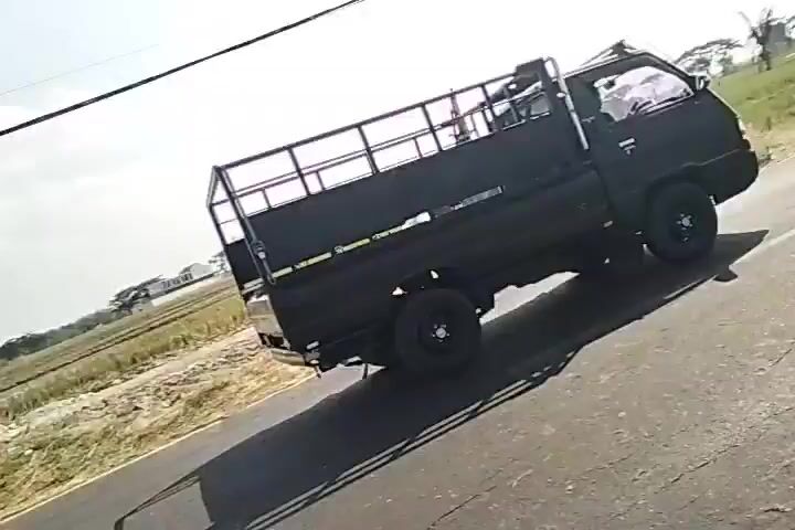 A black Mitsubishi pickup truck is seen driving down a rural road with fields and houses in the background. A motorbike carrying two passengers follows behind the truck. The sky is bright and overcast.