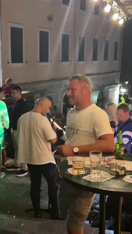 A man with short, blond hair and a white t-shirt is standing at a table outdoors, holding a cigarette to his mouth and smoking. He has a watch on his left wrist and is looking to his left. On the table in front of him are two glasses, one with a yellow liquid and ice, and another empty glass. There is also a small ashtray. Behind him and to his left, another man with a bald head is wearing a white t-shirt and facing away from the camera. Other people and buildings are visible in the background, suggesting a bar or restaurant setting at night.