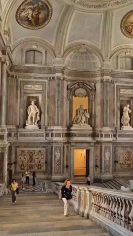The video shows a wide shot of the interior of a grand building, likely a palace or museum, with ornate marble walls and a high ceiling. In the foreground, a woman with blonde hair, wearing a black top and white pants, walks down a marble staircase. She is descending from right to left. In the background, there are several statues. On the left, a standing male figure in regal attire is in a niche. In the center, a seated female figure, wearing a crown and draped in cloth, is depicted with a lion beside her, also within a niche. Another male figure is visible in a niche on the far right. Several other people are seen standing and observing the art and architecture. The lighting is dim, with spotlights illuminating the statues.