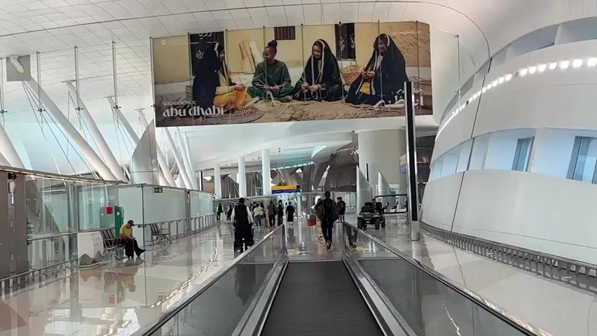 A wide shot captures an airport terminal with a large advertisement for "Experience Abu Dhabi" featuring four individuals in traditional attire. A moving walkway dominates the foreground, with a few travelers walking on the stationary floor to the left and right. More people are visible in the distance walking towards the advertisement. The terminal features high ceilings, modern architecture with white curved surfaces, and reflective flooring. The overall atmosphere appears to be calm and orderly.
