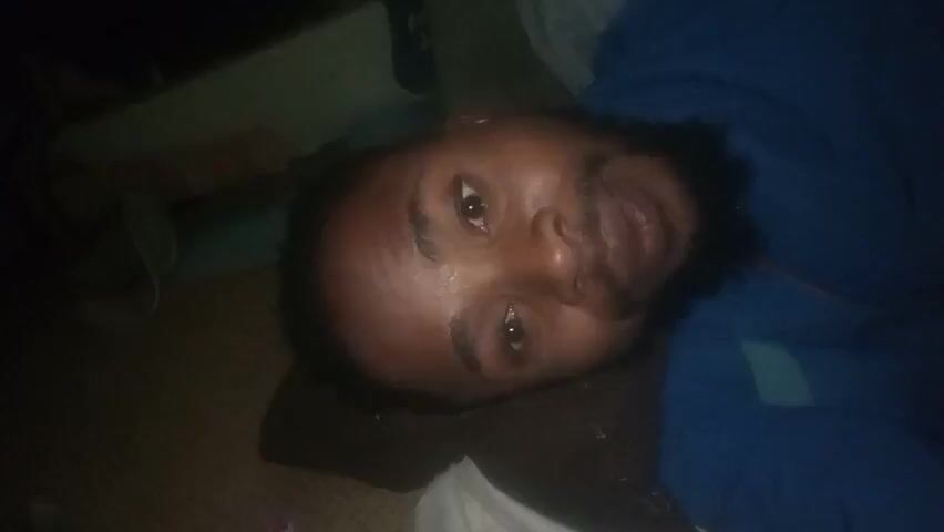 A man lies on his back, eyes partially closed and mouth open, with a bottle full of shelled nuts held near his face. The man is wearing a blue top. The environment is dimly lit, with furniture partially visible in the background. The camera angle is from above the man, looking down towards his face.