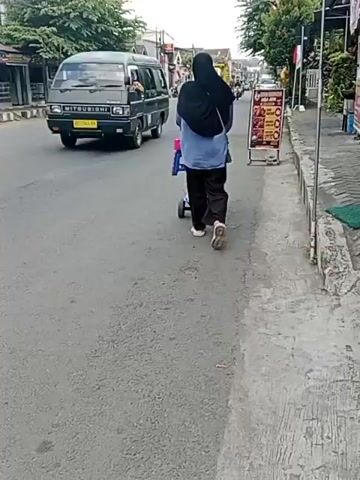 A person wearing a black hijab and dark pants pushes a stroller along a sidewalk. A minivan passes by on the left side of the road. A yellow dump truck is visible further down the road, followed by several other vehicles. A motorcycle rider is seen on the left side of the road. A sign with red text is placed on the sidewalk near the person. The environment appears to be a street in a town or city with buildings and trees lining the road. The weather seems to be overcast.