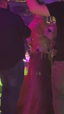 A woman is dancing or performing in a dimly lit club-like environment. The lighting changes between various colors, mostly blue, green, and yellow. She is wearing a decorative outfit consisting of a backless top and a long skirt that fades from pink to darker red. The top and skirt are adorned with floral and sparkly elements. The woman is surrounded by other people. In the background, there are tables with bottles of drinks on them. The camera is positioned behind the woman and remains mostly steady throughout the video.