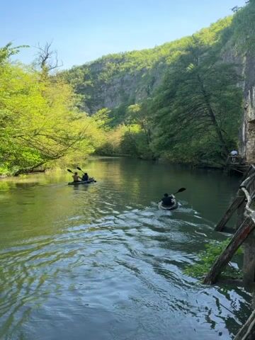 The video shows a tranquil river scene. Two kayaks are visible on the water; one contains two people, and the other has a single occupant who is paddling.  Adjacent to the river, a weathered wooden boardwalk follows the contours of a steep, rocky cliff face. A single individual is standing on a segment of the boardwalk, appearing to observe the water. Lush greenery, including trees and shrubs, surrounds the river and clifftop, completing the picturesque natural environment. The sky is bright with clear weather.